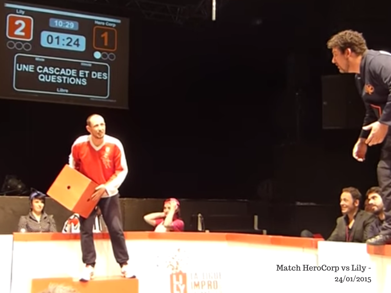 Capture d'écran de la vidéo suggérée dans l'article : on y voit 2 joueurs, un rouge et un bleu, sur la scène, ainsi que le panneau de scores et le titre de l'impro.